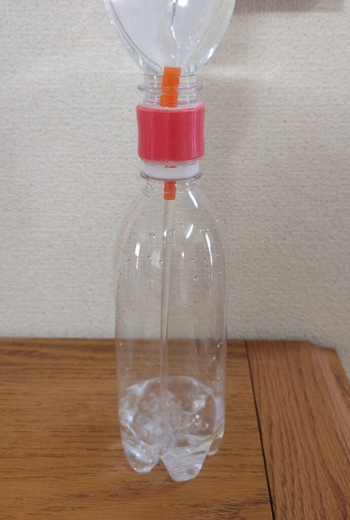 500mlのペットボトル2本を、赤い3Dプリント部品でつないだ水だけのタイマー。つなぎ目にオレンジのストローが通り、上のボトルの水が下のボトルに落ちている。