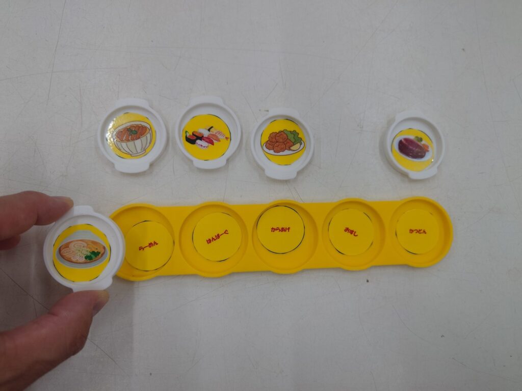 料理の絵が入った白い小皿の取っ手を指でつまみ、黄色いプレートにはめようとしている写真