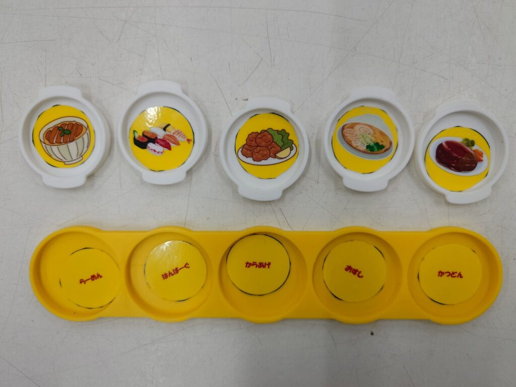 黄色いプレートの5つのくぼみにことばのラミネートを入れ、その上に料理の絵を入れた白い取っ手付き小皿を5つ並べた写真