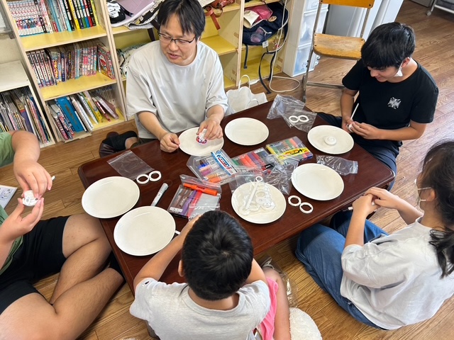 低い座卓を囲んで、子どもたちと大人がバトルコマの組み立てをしている写真。机の上には白い皿が6枚、袋に小分けした部品、色マジックのセットが並んでいる